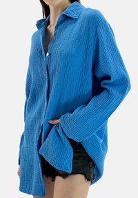 Blå skjorte med knapper lavet af tekstureret, krøllet stof. Har krave, lange ærmer og en afslappet pasform.