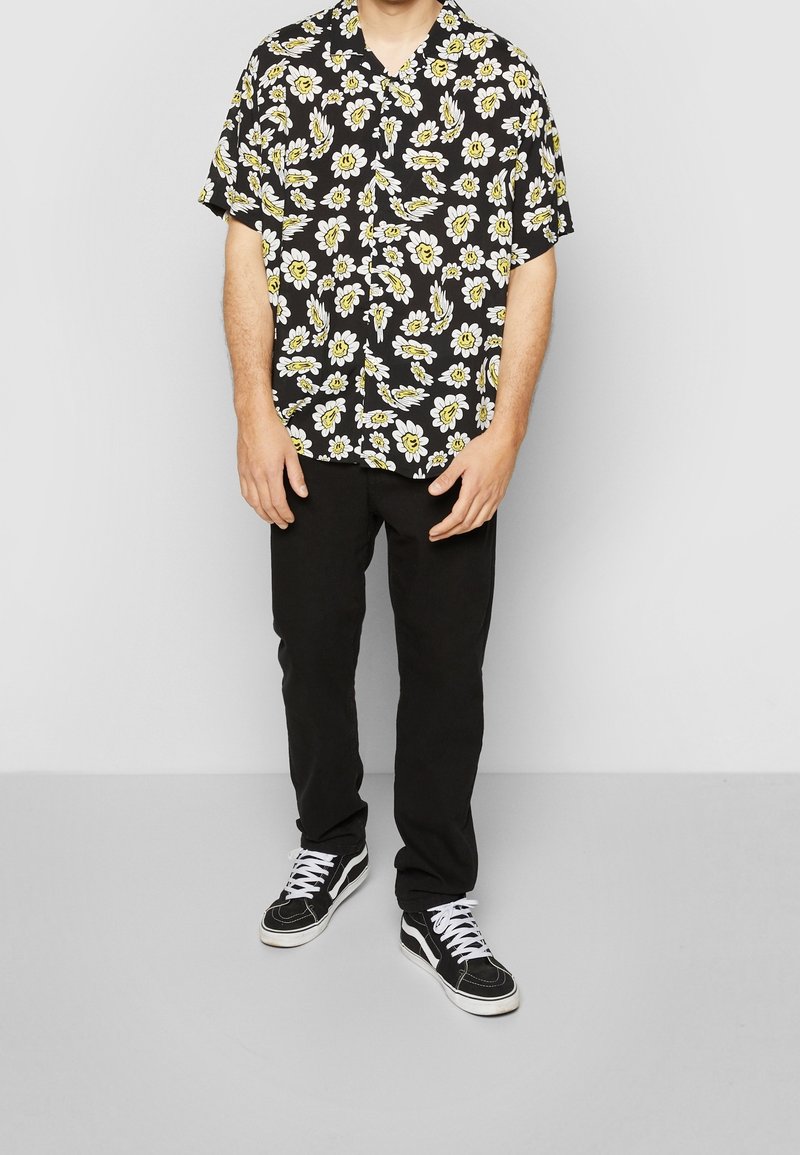 Chemise noire à manches courtes avec un motif floral jaune et blanc, associée à un pantalon noir et des baskets noires à lacets blancs.
