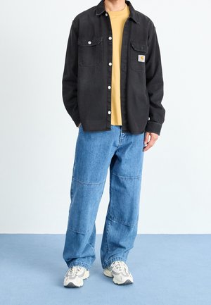 Camisa de mezclilla negra con dos bolsillos en el pecho, combinada con una camiseta amarilla mostaza y vaqueros azules sueltos, complementada con zapatillas beige.