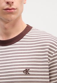 Brązowa i biała bluzka w paski z żebrowanym brązowym kołnierzykiem, z małym brązowym logo "CK" na piersi. Teksturowana bawełniana tkanina.