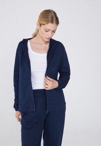 Sudadera con cremallera en color marino, con un bolsillo frontal, capucha con cordón y puños de ribete, utilizada sobre una camiseta sin mangas blanca y combinada con pantalones a juego.