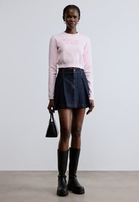 Maglione rosa corto con logo, abbinato a una minigonna di denim scuro con pieghe e bottone. Stivali neri con plateau fino al ginocchio e borsa.