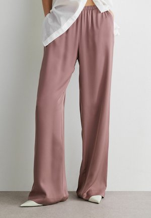 Person trägt lockere satinartige Hose in Mauve mit elastischem Bund, kombiniert mit weißen spitzen Schuhen und einem weißen transparenten Oberteil.