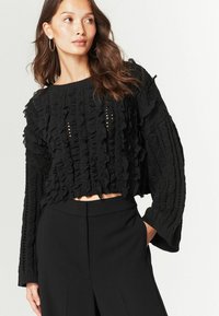 Fekete cropped pulóver, texturált anyaggal, fodrokkal és nyitott mintázatú részletekkel. Hosszú ujjú, kontrasztos kötött mintával.