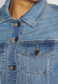 Vaaleansininen farkkutakki, jossa on kaulus, mustat napit ja kaksi rintataskua. Huomiota herättävä kädenjälki ja vaaleanruskeat kontrastitikkaukset.