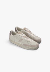 Pareja de zapatillas bajas de ante beige con cordones blancos, suelas de goma texturizadas y logotipo de calavera y huesos cruzados en el lateral.