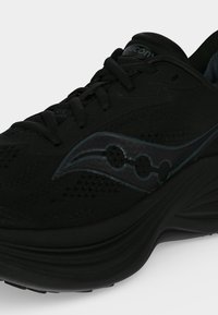 Μαύρο αθλητικό παπούτσι Saucony με παχιά σόλα, δετό σχέδιο και λογότυπο στο πλάι, εμφανίζεται από πλάγια και μπροστινή γωνία.