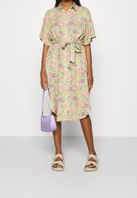 Vestido floral con mangas cortas, ceñido en la cintura, con estampados en verde, rosa y púrpura. Combinado con un bolso lavanda y sandalias.