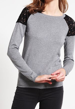 Vrouw draagt een grijze trui met lange mouwen met zwarte kantdetails op de schouders, gecombineerd met een zwarte broek, handen zachtjes samengevouwen.