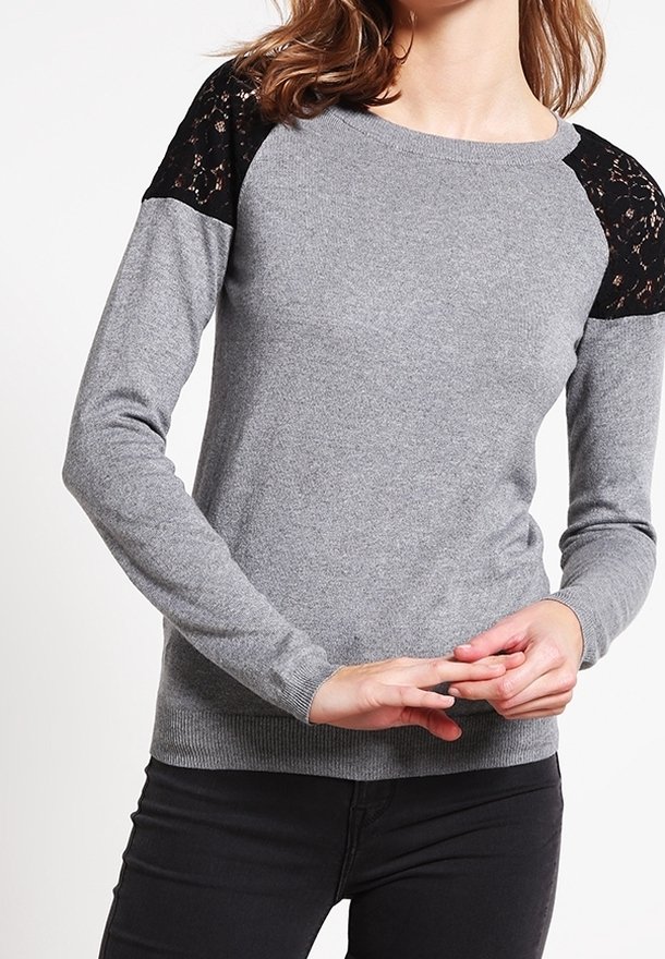 Femme portant un pull gris à manches longues avec des détails en dentelle noire aux épaules, associé à un pantalon noir, mains doucement jointes.