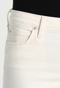 Jean en denim blanc avec un design classique à cinq poches, doté d'une fermeture à bouton métallique et de coutures visibles. Texture douce avec une légère élasticité.