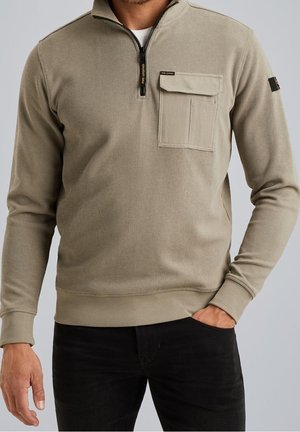 Mężczyzna noszący beżowy sweter z suwakiem do ćwiartki i kieszenią na piersi oraz czarne spodnie, ręka w kieszeni, na jednolitym jasnym tle.