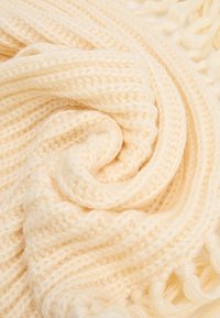 Gehaakte crèmekleurige sjaal, zachte textuur, met een gevlochten patroon en franjes aan de randen, opgerold in een spiraal ter display.