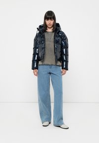 Marineblauwe gewatteerde jas met een glanzende afwerking, met ritssluiting aan de voorkant en capuchon, gedragen over een grijze sweatshirt en lichtblauwe wijde jeans.