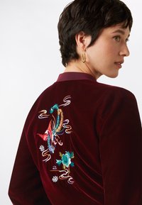 Giacca in velluto bordeaux con ricamo di drago e fiori sul retro. Presenta un colletto e un materiale testurizzato, evidenziando dettagli intricati.