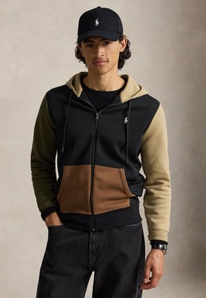 Jeune homme portant une casquette noire et un sweat à capuche à blocs de couleur avec des manches noires, marron et beige, debout devant un fond beige uni.