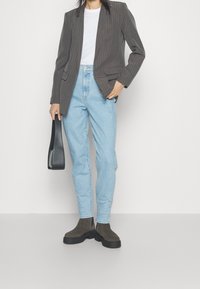 Donna che indossa un blazer grigio a righe, una camicia bianca, jeans azzurri chiari e stivali scuri, mentre tiene una borsa nera su uno sfondo neutro.
