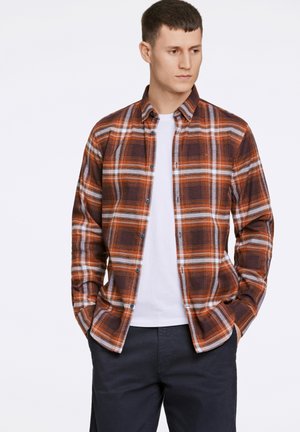 Camisa de cuadros en colores naranja, marrón y blanco con diseño de botones. Debajo lleva una camiseta blanca, combinada con pantalones oscuros.