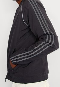 Black lightweight jacket with long sleeves featuring grey striped accents. Includes side pockets and elasticated waistband for fit.