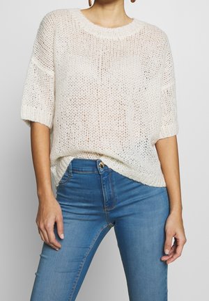 Femme portant un pull blanc à manches courtes à maille lâche, rentré dans un jean bleu taille haute, debout les mains détendues le long du corps.