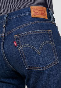 Denim jeans in een diepe blauwe kleur, stevige stof, onderscheidende stiksels, een leren logopatch en een kenmerkende rode tab op de achterzak.