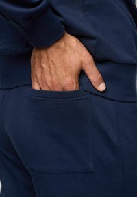 Granatowe dresy z tkaniny z boczną kieszenią. Materiał wydaje się miękki w dotyku, o gładkiej fakturze, zaprojektowane z myślą o komforcie i codziennym noszeniu.