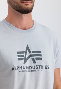 Helles graues Baumwoll-T-Shirt mit einem schwarzen Grafik-Logo, das Sterne und Streifen zeigt, sowie dem Text "ALPHA INDUSTRIES, KNOXVILLE, TENNESSEE, U.S.A.".