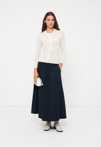 Light ribbed knit cardigan in cream, paired with a navy pleated maxi skirt. Accessories include a small white handbag and light grey slip-on shoes.