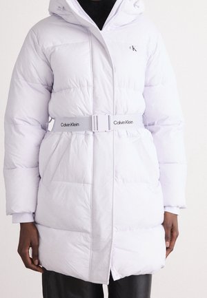 Manteau matelassé blanc avec capuche, ceinture Calvin Klein à la taille, porté sur un top et un pantalon noirs, mains détendues sur les côtés.