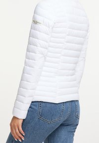 Vit puffjacka med horisontellt stitchad stoppning, rundade axlar och en något avsmalnande passform. Slät textur, minimal logotyp på ärmen.