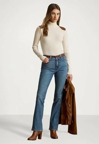 Moteris, vilkinti kreminę marškinių golfą su pečių detalėmis, mėlynas džinsas ir rudos aukštakulnės batai, viena ranka laikanti rudą odinį švarką.