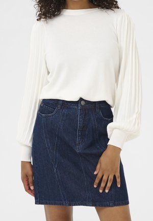 Femme portant un chemisier blanc à manches longues plissé, rentré dans une jupe en denim bleu foncé taille haute avec des poches à l'avant.