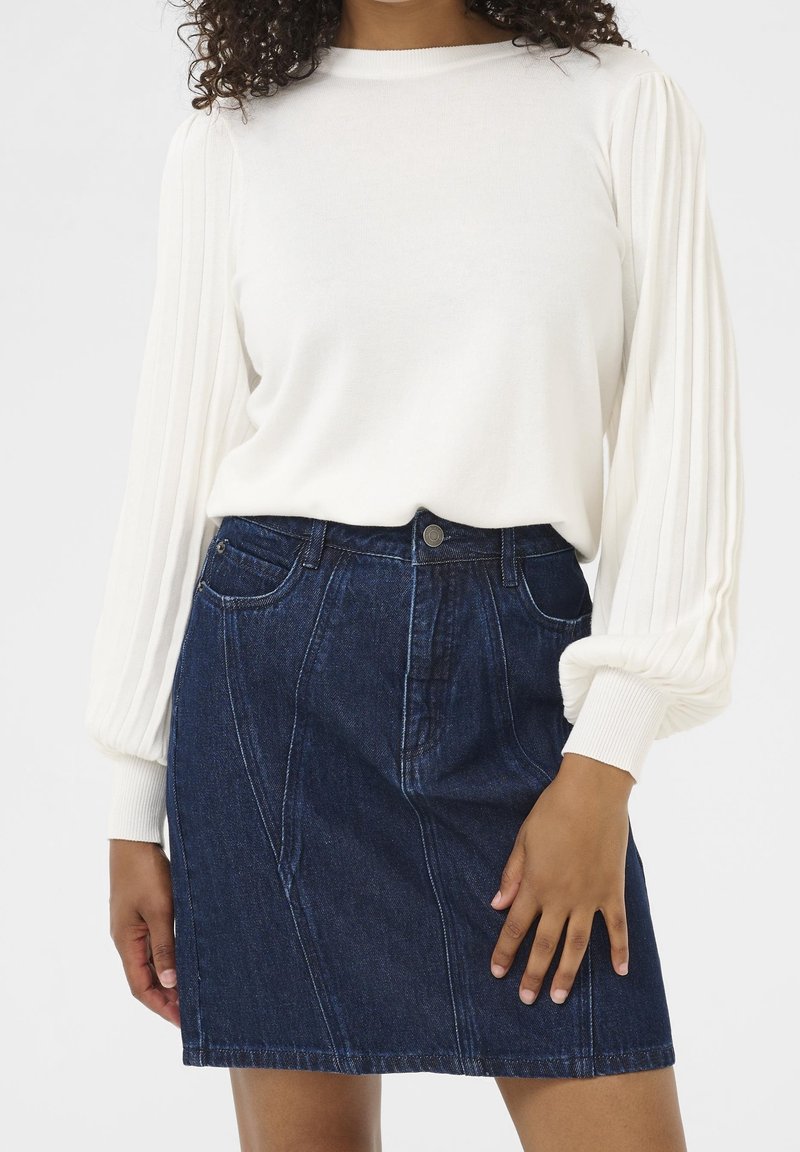 Femme portant un chemisier blanc à manches longues plissé, rentré dans une jupe en denim bleu foncé taille haute avec des poches à l'avant.
