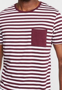 Camiseta a rayas rojas y blancas con cuello redondo y un bolsillo en el pecho. La tela parece suave con una textura lisa. Mangas cortas.