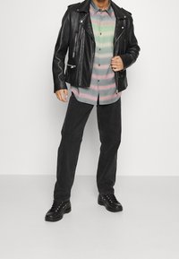 Chaqueta de motorista de cuero negro sobre una camisa de botones con rayas multicolores. Pantalones oscuros y botas negras con cordones completan el conjunto.