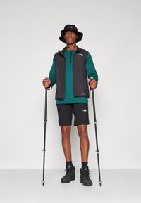 Une personne en gilet noir sur une chemise à manches longues verte, portant un short noir et des chaussures de randonnée, tenant des bâtons de trekking et portant un chapeau de randonnée noir.