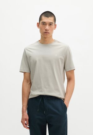 Junger Mann mit kurzen Haaren, der ein hellgraues kurzärmeliges T-Shirt und dunkelblaue Kordelhose trägt, steht vor einem schlichten Hintergrund.