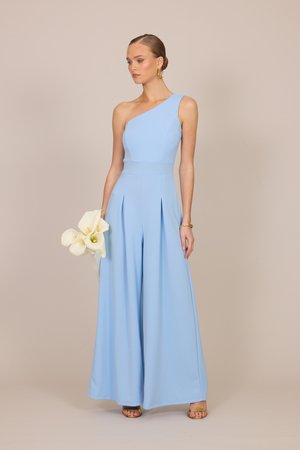 Nő világoskék egyrészes, váll nélküli ruhában fehér kalla liliomokat tart, arany karika fülbevalót és arany szandált visel bézs háttér előtt.