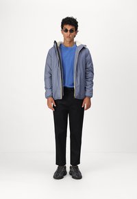 Giacca a vento grigia con cappuccio foderato in bianco, indossata sopra una camicia blu. Pantaloni neri affusolati e sneakers grigie completano l'outfit.