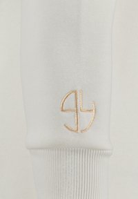 Manica di un felpa bianca con polsino a coste, caratterizzata da un logo ricamato dorato, che mette in mostra una superficie di tessuto liscia e testurizzata.