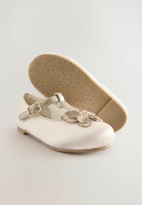 Weiße Satin-Kinderschuhe mit einem silbernen Riemen, einer dekorativen metallischen Schmetterlingsapplikation auf der Vorderseite und einer gemusterten Gummisohle.