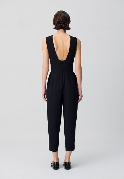 Combinaison sans manches noire avec un dos en V profond, jambes fuselées et fermeture éclair. Fabriquée dans un tissu lisse et mat.