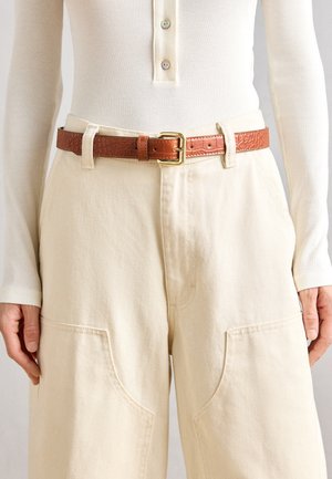 Brūna teksturēta ādas josta ar zelta taisnstūra sprādzi, uzvilkta virs krēmkrāsas, brīva griezuma biksēm ar lieliem priekšējām kabatām.