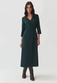 Robe enveloppante vert foncé avec des manches trois-quarts, une taille cintrée et une jupe longue, fabriquée à partir d'un tissu doux et texturé.