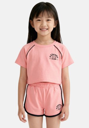 Jeune fille souriante, portant un crop top manches courtes rose ellesse CLUB et un short assorti avec une bordure noire sur fond blanc.
