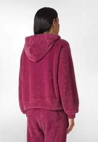 Felpa fuzzy con cappuccio di colore rosa scuro, con un design corto, polsini a costine e una texture morbida. Si abbina ai pantaloni coordinati.