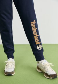 Námornícke tepláky sú zdobené zlatým textom a logom "Timberland" po celej nohe, v kombinácii s béžovými a hnedými športovými teniskami.