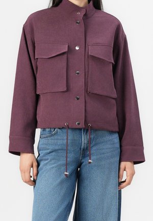 Kobieta w śliwkowej kurtce z zatrzaskami, dwoma dużymi przednimi kieszeniami na klapę oraz regulowanymi sznurkami u dołu, w połączeniu z niebieskimi dżinsami.