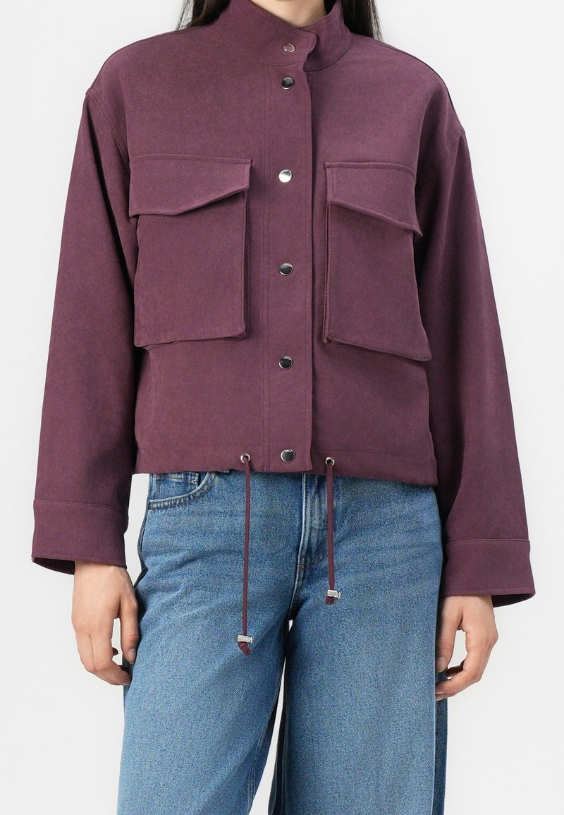 Femme portant une veste couleur prune avec des boutons-pression, deux grandes poches avant à rabat, et des cordons ajustables à l'ourlet, associée à un jean bleu.