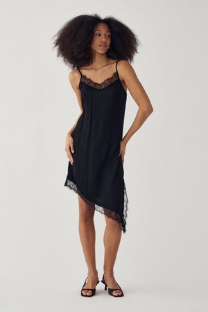 Frau mit natürlichen lockigen Haaren, die ein asymmetrisches schwarzes Slip-Kleid mit Spitzenbesatz und schwarze offene Absätze trägt, stehend vor einem schlichten Hintergrund.